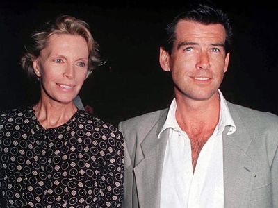 Pierce Brosnan is wearing a grey suit and a white shirt and Cassandra Harris is wearing a black dress with some designs on it.
